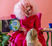 A drag queen, dressed all in pink, holds up a book while interacting with kids during a story time reading event