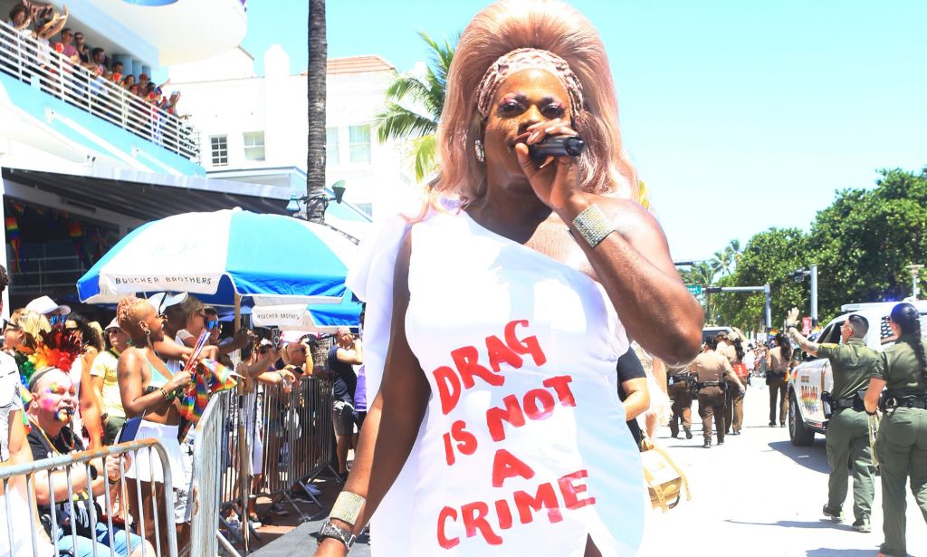 A Black drag performer speaks into a microphone during an LGBTQ+ event in Florida while wearing a white dress with red lettering that reads: 'Drag is not a crime'