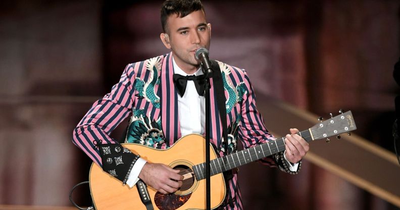 Sufjan Stevens wears a pink and black striped suit jacket as he plays the guitar and sings into a microphone on stage