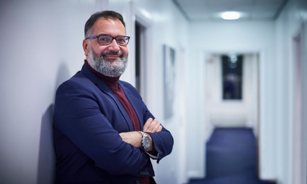 Ioannis Ntanos, one of the leading surgeons on trans healthcare in the UK, wears a turtleneck and suit jacket as he leans on a wall with his arms crossed