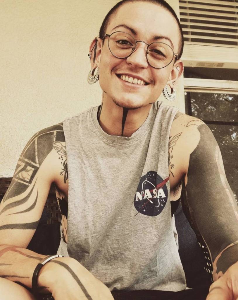 Avery Wade Kelley, a trans person from Florida with a variety of tattoos and glasses, wears a sleeveless shirt with the NASA logo on it as he sits outside his house