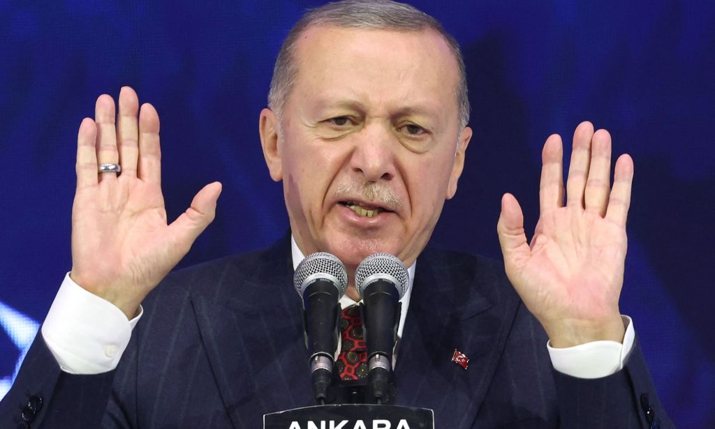 Turkey's president Recep Tayyip Erdoğan, who has a history of making anti-LGBTQ+ remarks, wears a suit and tie as he holds up both his hands while speaking into a microphone