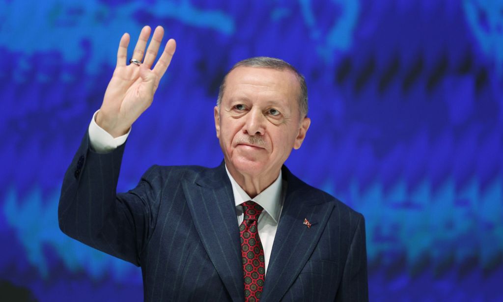 Turkey's president Recep Tayyip Erdoğan, who has a history of making anti-LGBTQ+ remarks, wears a suit and tie as he holds up one hand during a political conference