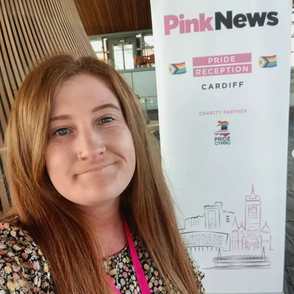 This is an image of a woman standing in front of a banner that says "PinkNews" at the top. She s smiling and has long red hair.