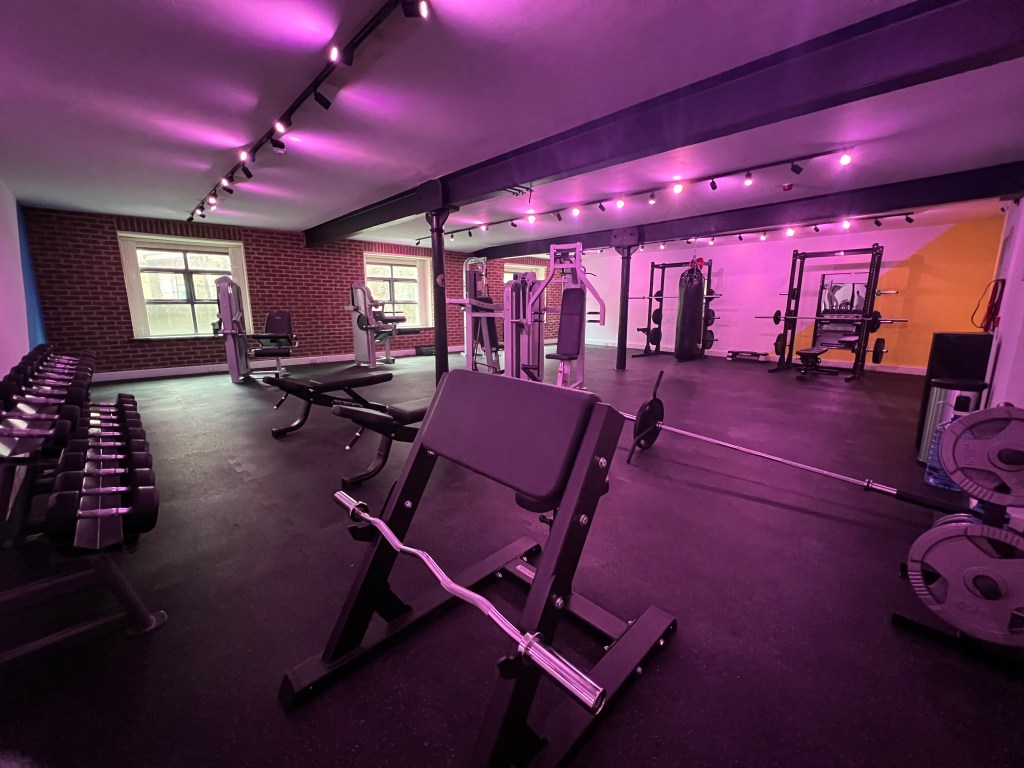 A picture of the inside of the Leeds People's Gym, which was set up by Daniel Browne and Chris Woods. It shows purple hued mood lighting in the weights room.