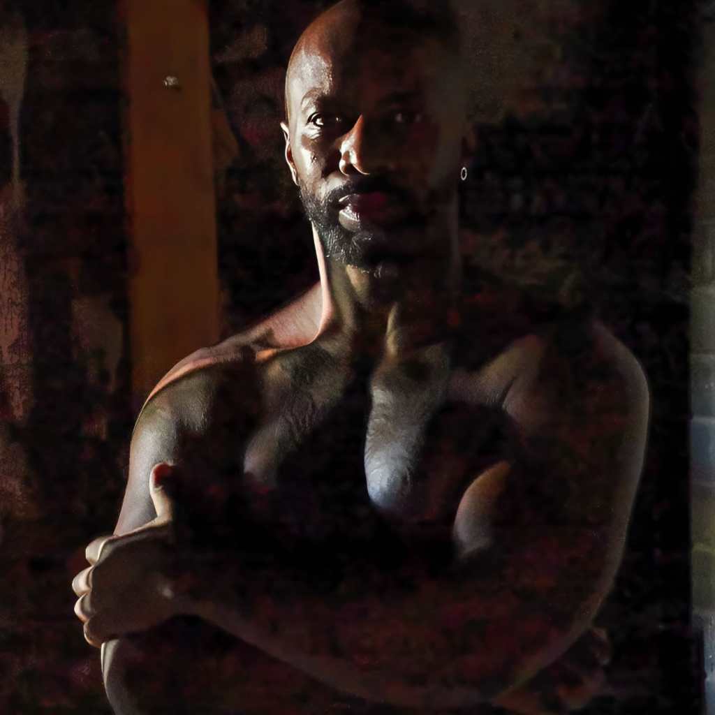 Race Cooper pictured shirtless folding his arms in a dark room.
