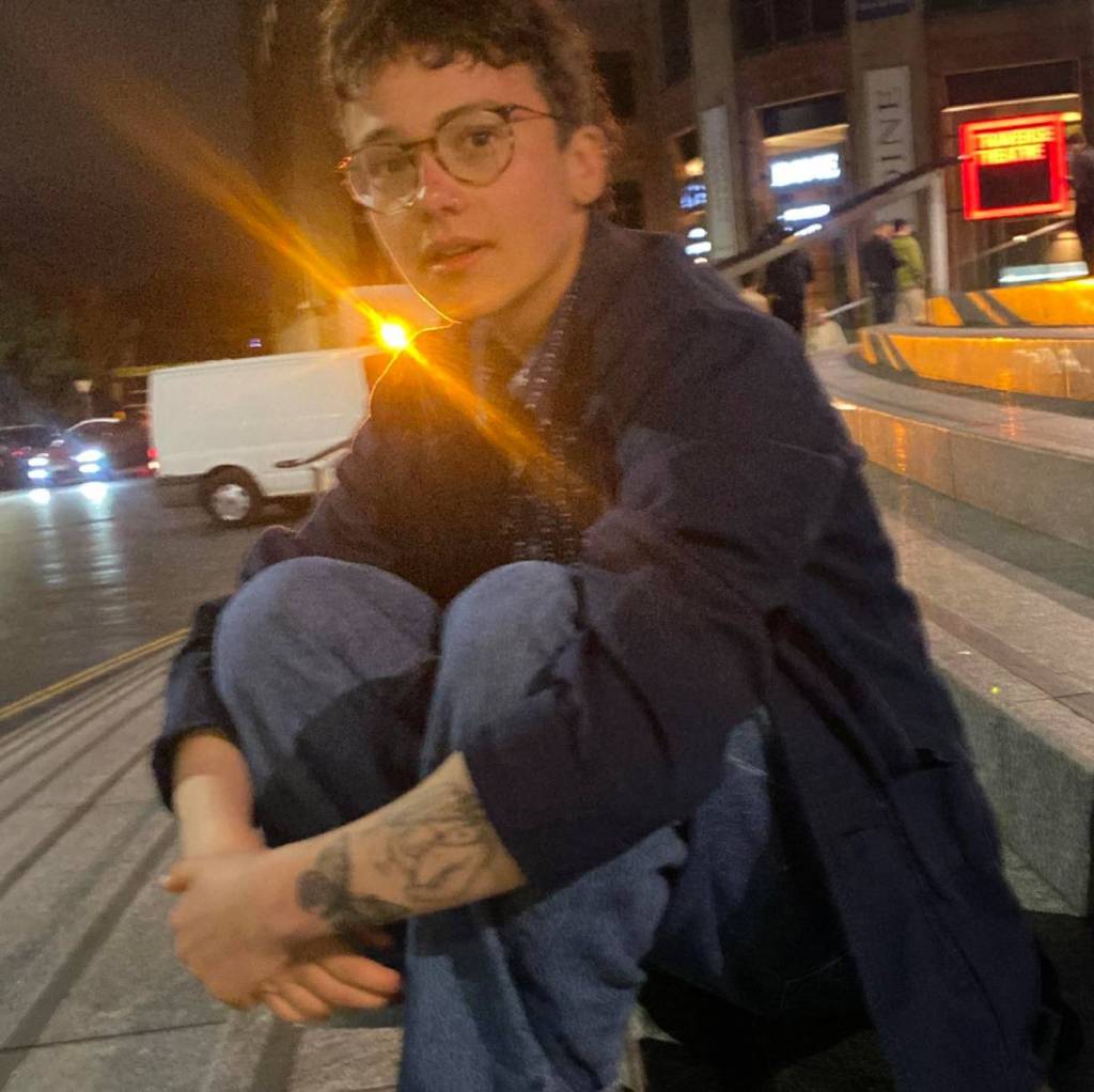 Trans, non-binary person Mizy, who has had top surgery, wears jeans and a dark top as they pose while sitting on some stairs