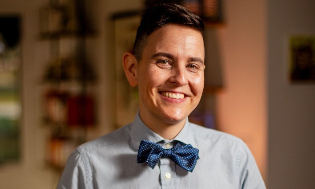 Dr Erin Baker, a non-binary person who had top surgery, smiles while wearing a button up shirt and bow tie