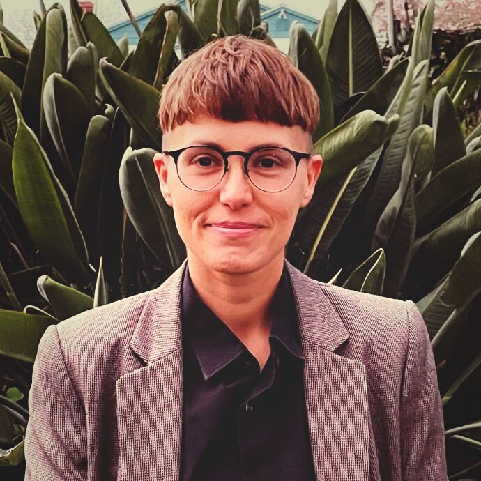 A picture of AJ Schaerer, a trans person, wearing a button up shirt and suit jacket as he stands in front of a green plant