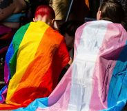 Two people sit together wearing a trans flag and a Pride flag as capes.