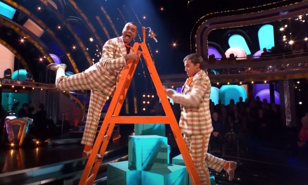 Layton Williams and Nikita Kuzmin wear matching plaid suits as they stand on a ladder during a performance on BBC's Strictly Come Dancing