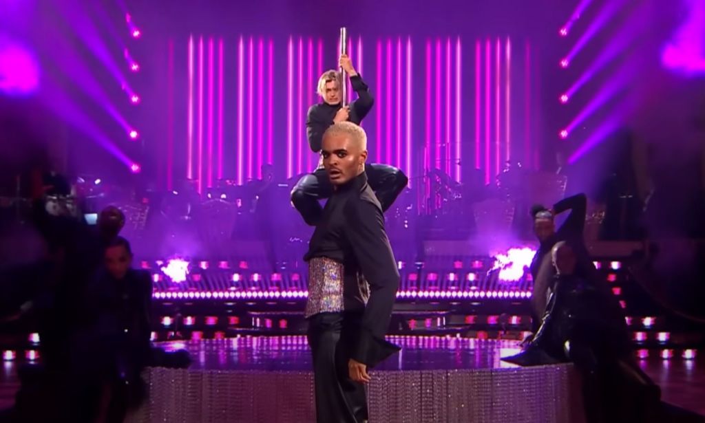 Layton Williams and Nikita Kuzmin wear matching sparkly corsets and black outfits as Williams stands in front of Kuzmin, who is clutching a pole, during a performance on BBC's Strictly Come Dancing