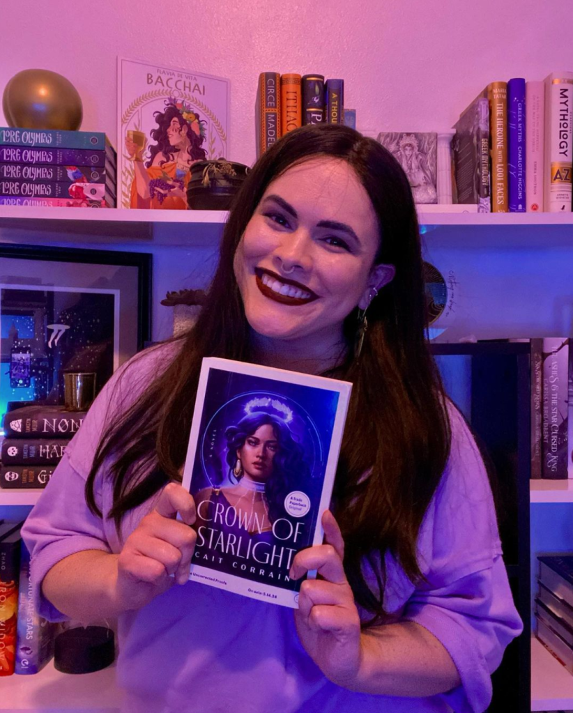 Author Cait Corrain wears a jumper as they hold up a cover for their book in pinky-blue lighting