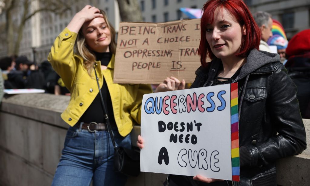 Two people stand side by side during an LGBTQ+ rights protest to ban conversion therapy. One person holds up a sign reading 'queerness doesn't need a cure'