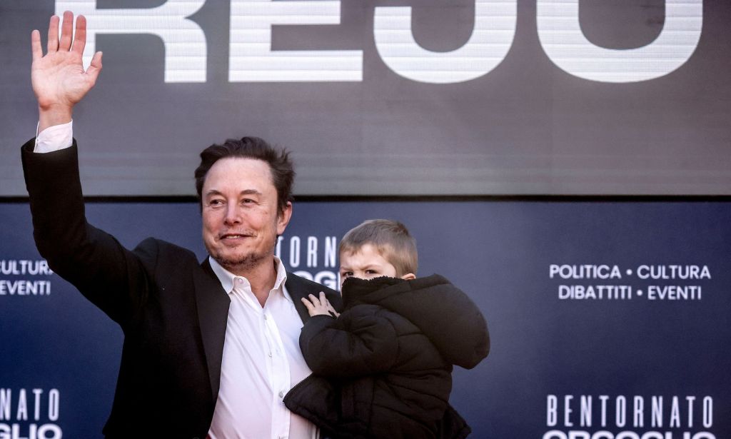 Elon Musk waves to people off camera as he holds one of his children in his other arm. At the right-wing event, Musk claimed people should have more babies