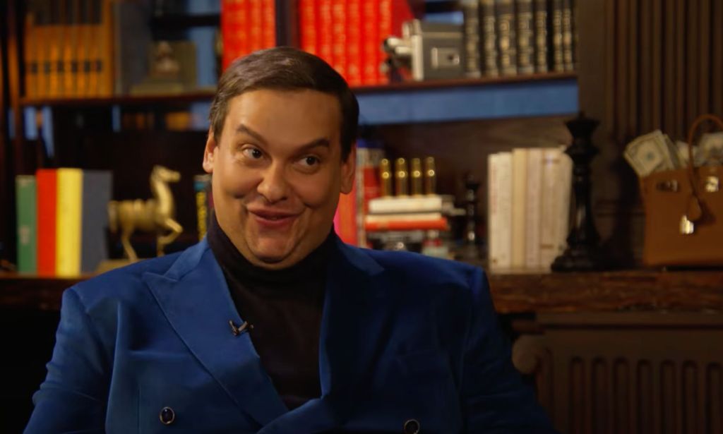 George Santos wears a black shirt and blue jacket as he sits in front of a bunch of books during an interview with Ziwe. In this frame, he is making a funny face