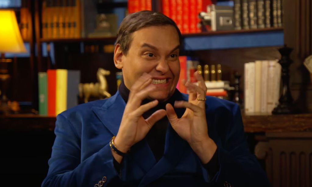 George Santos wears a black shirt and blue jacket as he sits in front of a bunch of books during an interview with Ziwe. In this frame, he is smiling as he gestures with his hands in front of his face