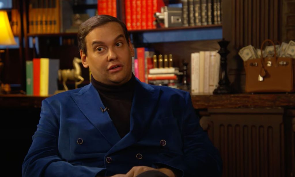 George Santos wears a black shirt and blue jacket as he sits in front of a bunch of books during an interview with Ziwe. In this frame, he's looking off to the side
