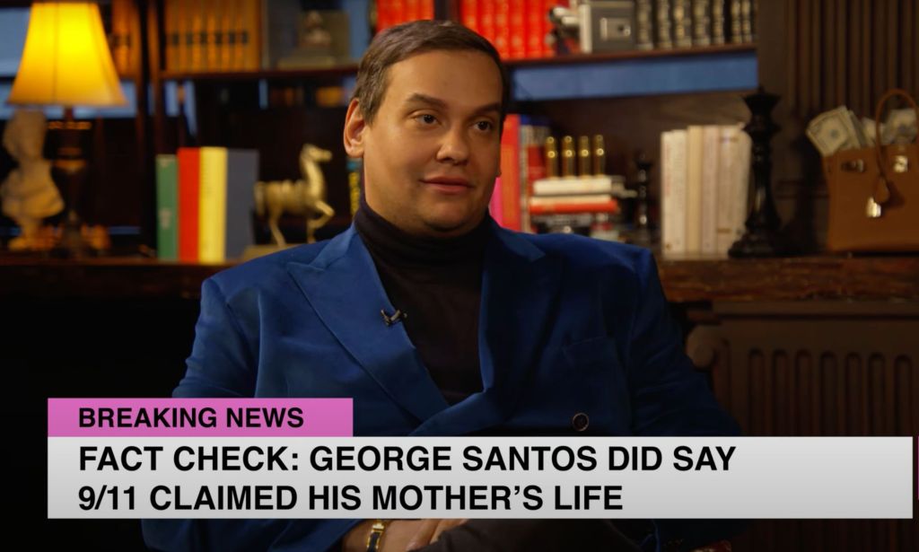 George Santos wears a black shirt and blue jacket as he sits in front of a bunch of books during an interview with Ziwe. In this frame, there's a little line that says "Fact check: George Santos did say 9/11 claimed his mother's life."