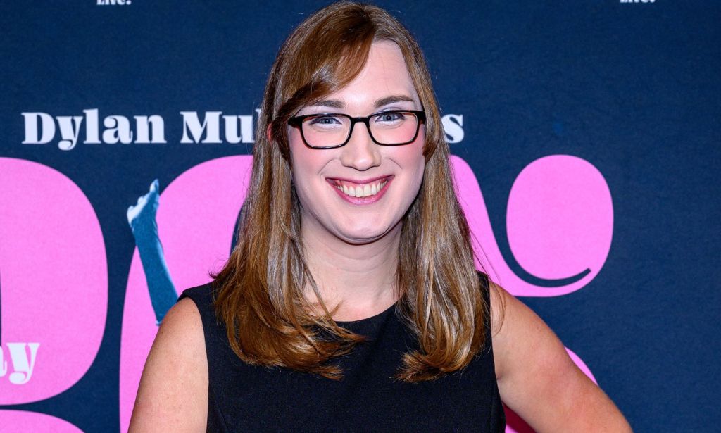 Delaware senator Sarah McBride wears a black dress as she smiles for the camera in front of a pink and blue background