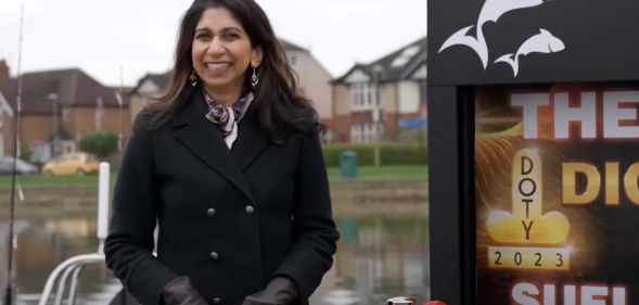 Former home secretary Suella Braverman, wearing a black coat, smiles awkwardly, as she's presented with The Last Leg's "D**k of the Year" award which is displayed on a sign behind her