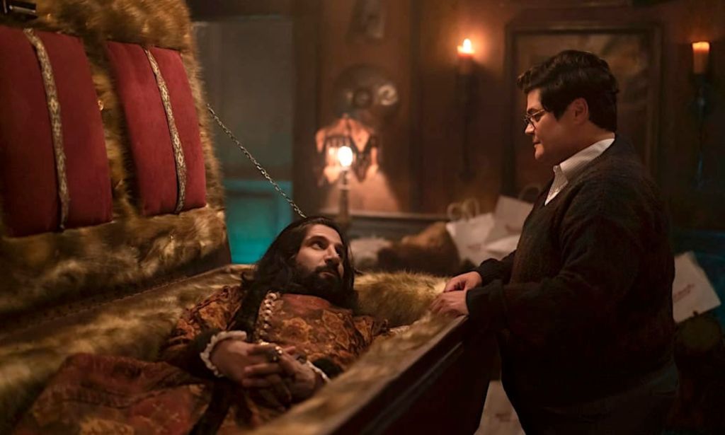 A still from What We Do in the Shadows with vampire character Nandor in a coffin with human familiar Guillermo talking to him