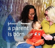 This is an image with 2 women holding a baby. The woman on the left is wearing a grey jumper. She has long dark hair and is wearing glasses. The woman on the right has bright blonde hair and is wearing sunglasses. The baby in the middle is wearing an orange onesie. Over the image in white text reads "JOHNSON'S, PINKNEWS A PARENT IS BORN"