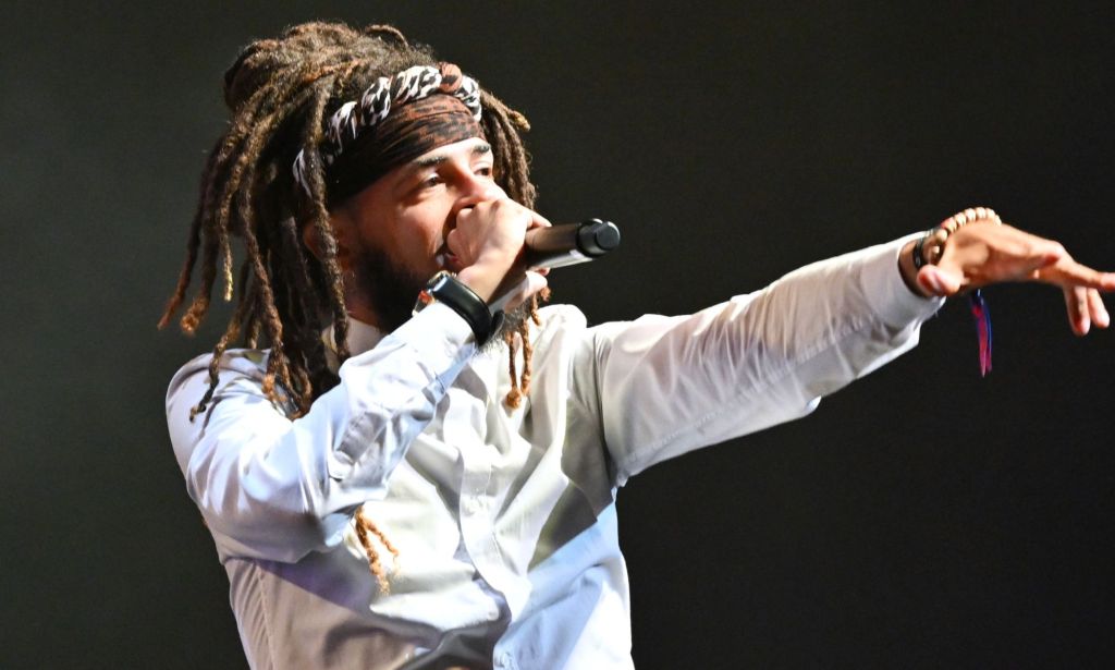 A man with dreadlocks and a bandana sings into a microphone and dances.