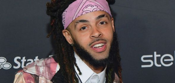 A man with dreadlocks and a chin strap beard wearing a pink bandana poses during a red carpet event.