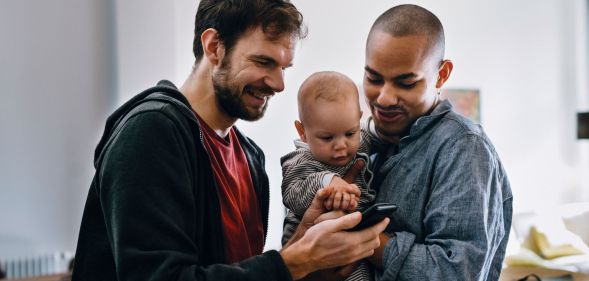 Two men holding a baby watch a video on their mobile phone.