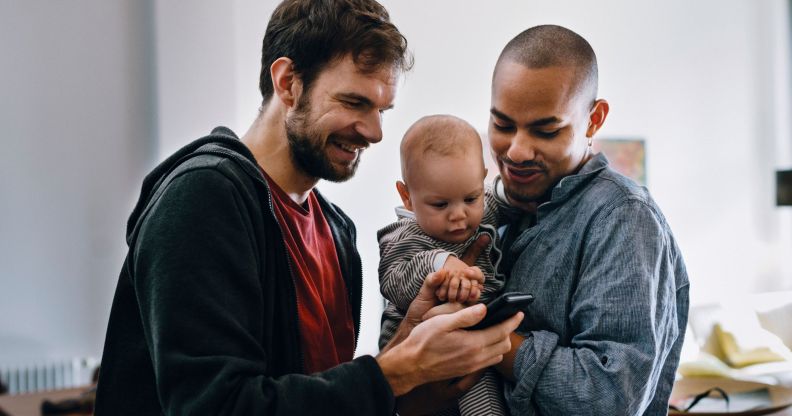Two men holding a baby watch a video on their mobile phone.