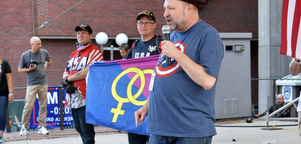 Image shows three men on a stage, the middle man is holding a straight pride flag that shows the symbols for man and woman on a blue and pink background