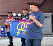 Image shows three men on a stage, the middle man is holding a straight pride flag that shows the symbols for man and woman on a blue and pink background