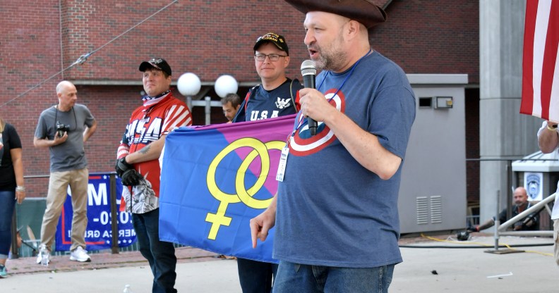 Image shows three men on a stage, the middle man is holding a straight pride flag that shows the symbols for man and woman on a blue and pink background