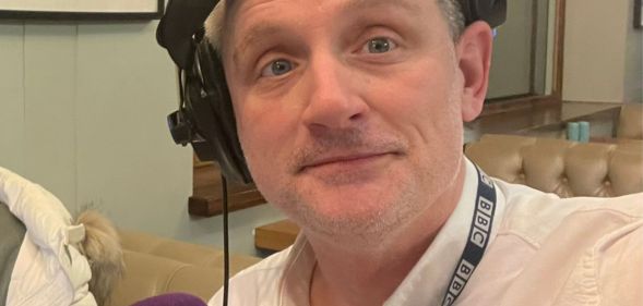 A man in a white button shirt wearing a pair of headphones on one ear smiles.