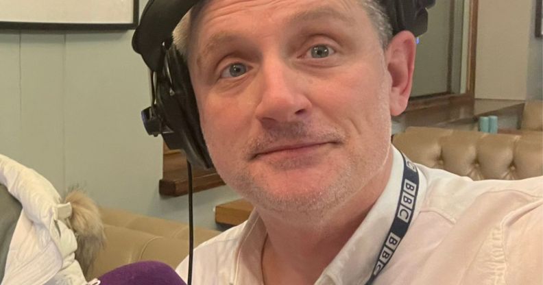 A man in a white button shirt wearing a pair of headphones on one ear smiles.