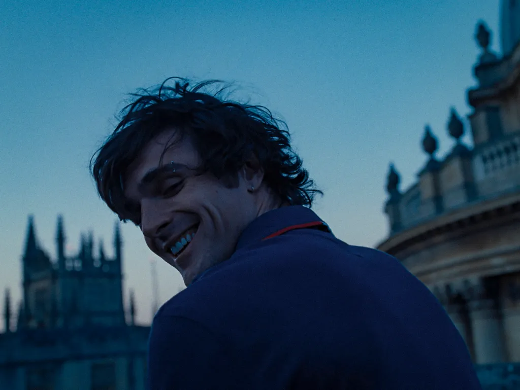 Jacob Elordi as Felix in Saltburn, it's twilight and Felix's is turning his head towards the camera, smiling.