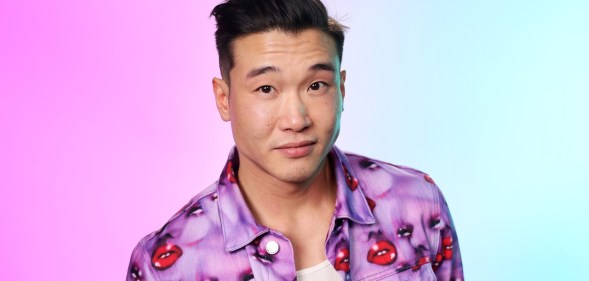 Joel Kim Booster in a jacket with purple faces and red lips. The background is hazy pink and blue.