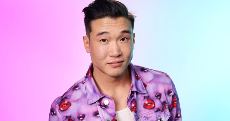 Joel Kim Booster in a jacket with purple faces and red lips. The background is hazy pink and blue.