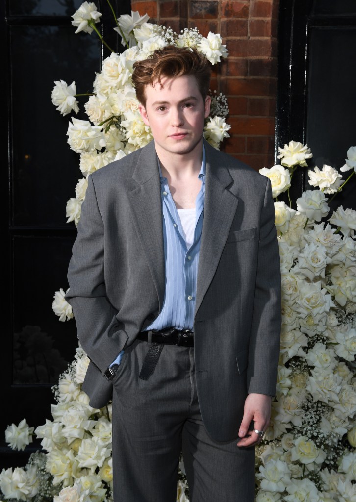 Kit Connor in a grey suit in front of a white flower display.