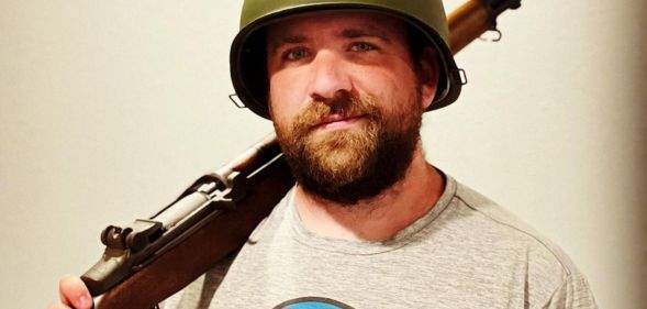A man wearing a green army helmet holds a wooden rifle over his shoulder.