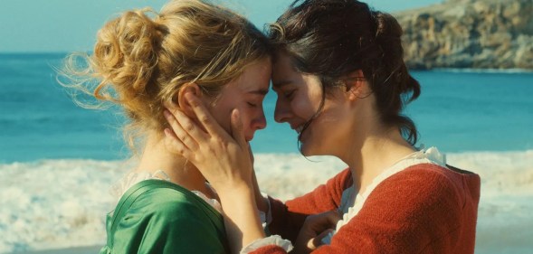 Portrait of a Lady on Fire still: two women, one blonde in a green dress, one brunette in a red gress, are clutching eachother's faces.