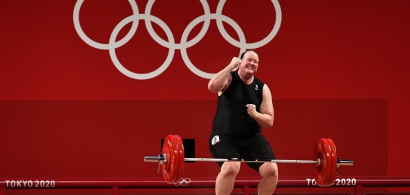 Weightlifter Laurel Hubbard is a trans woman. She's celebrating after lifting a weighted bar. Red backround with Tokyo 2020 Olympic Games branding.