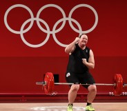 Weightlifter Laurel Hubbard is a trans woman. She's celebrating after lifting a weighted bar. Red backround with Tokyo 2020 Olympic Games branding.