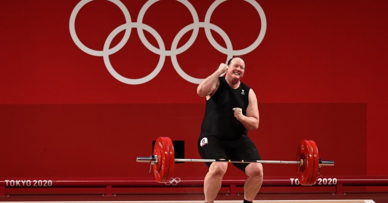 Weightlifter Laurel Hubbard is a trans woman. She's celebrating after lifting a weighted bar. Red backround with Tokyo 2020 Olympic Games branding.