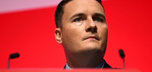 A politician at a podium with two microphones stands infront of a red screen.