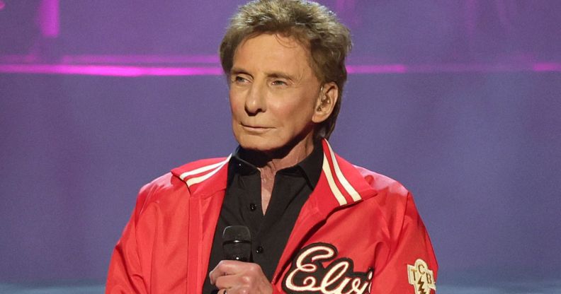 Singer Barry Manilow wears a black shirt and red jacket as he holds a microphone in front of his chest