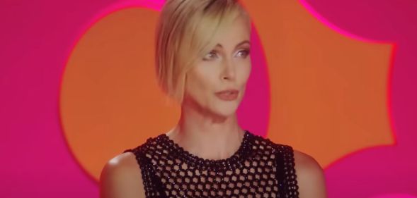 Charlize Theron wears a black outfit as she sits in front of a pink and orange background as a guest judge on Drag Race