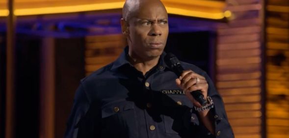 Dave Chappelle, who has faced backlash for making anti-trans jokes, wears a dark shirt as he speaks into a microphone in his new Netflix special The Dreamer