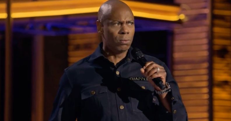 Dave Chappelle, who has faced backlash for making anti-trans jokes, wears a dark shirt as he speaks into a microphone in his new Netflix special The Dreamer